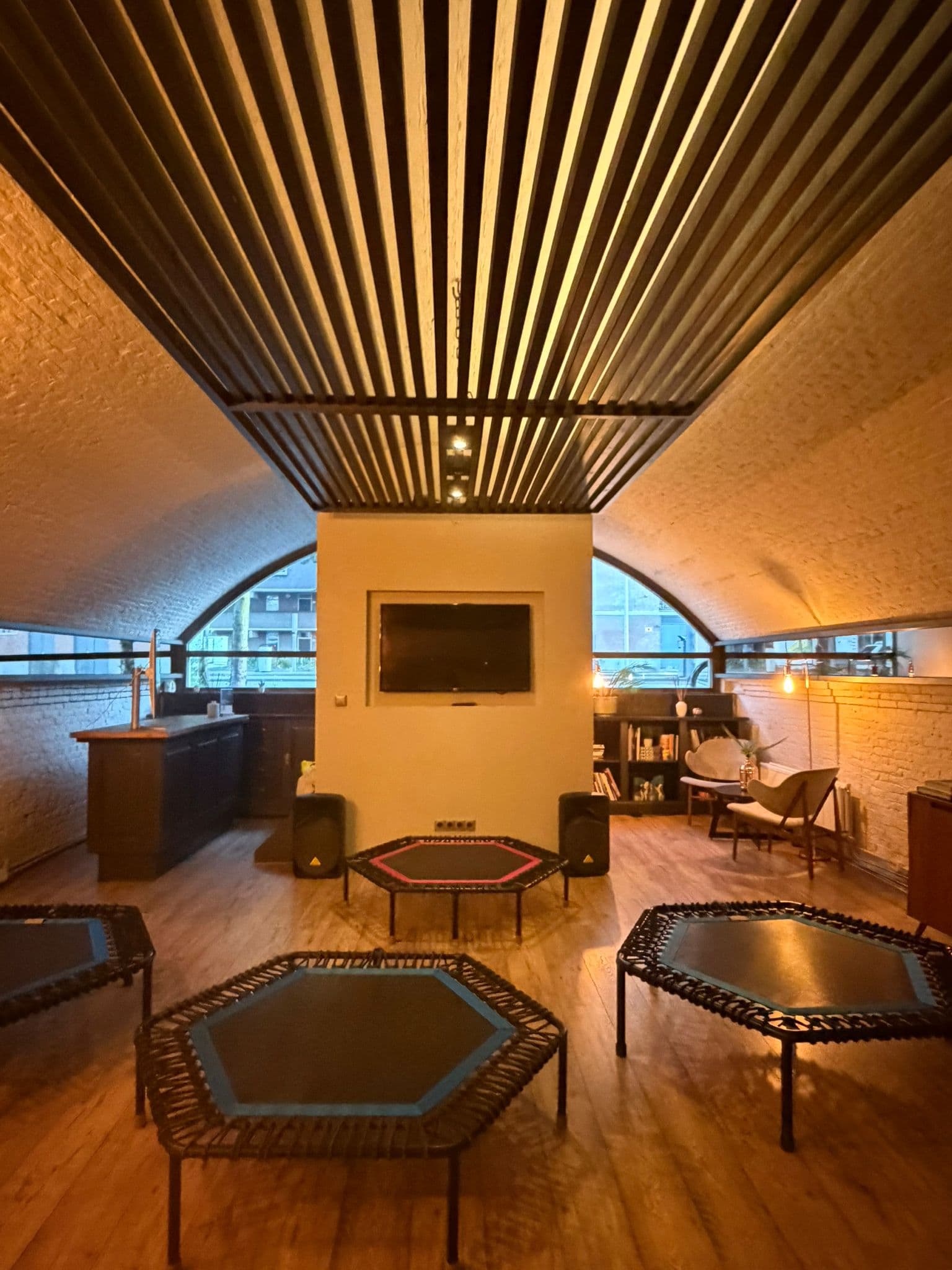 The arch interior looking toward the far windows — a whitewashed vaulted brick ceiling, a dark slatted ceiling panel, a wooden floor, trampolines set out, a bar counter on one side and armchairs on the other