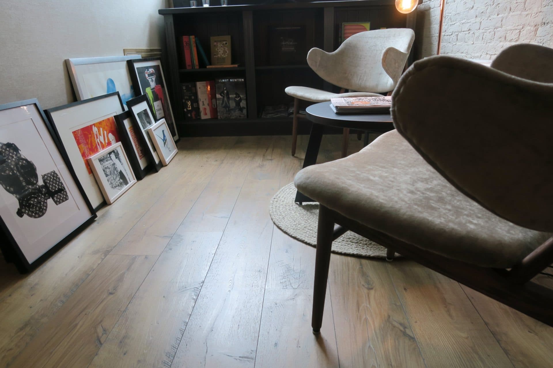 Ingelijste prints tegen de muur naast een donkere boekenkast met kunst- en fotoboeken, en een gestoffeerde loungestoel op de brede houten vloerdelen