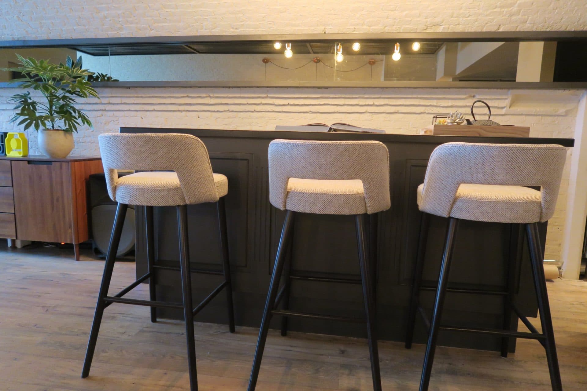 Drie gestoffeerde barkrukken met zwarte poten aan een donkere bar met paneelwerk, tegen witgekalkt metselwerk met een spiegelstrook waarin de hanglampen weerspiegelen