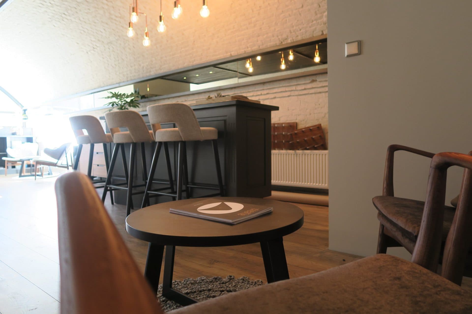 Zicht vanuit de loungestoelen over een ronde salontafel naar de bar en de barkrukken, met koperen hanglampen aan het bakstenen plafond