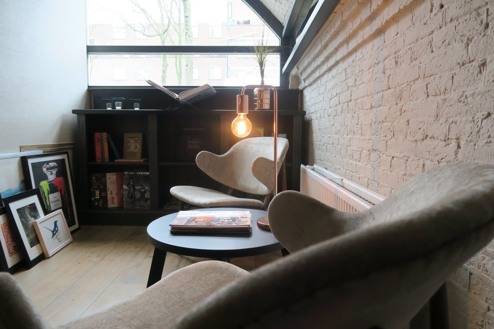 De leeshoek bij het raam: twee crèmekleurige loungestoelen rond een zwart bijzettafeltje met tijdschriften, een koperen staande lamp, een boekenkast met kunstboeken en ingelijste prints tegen het witgekalkte metselwerk