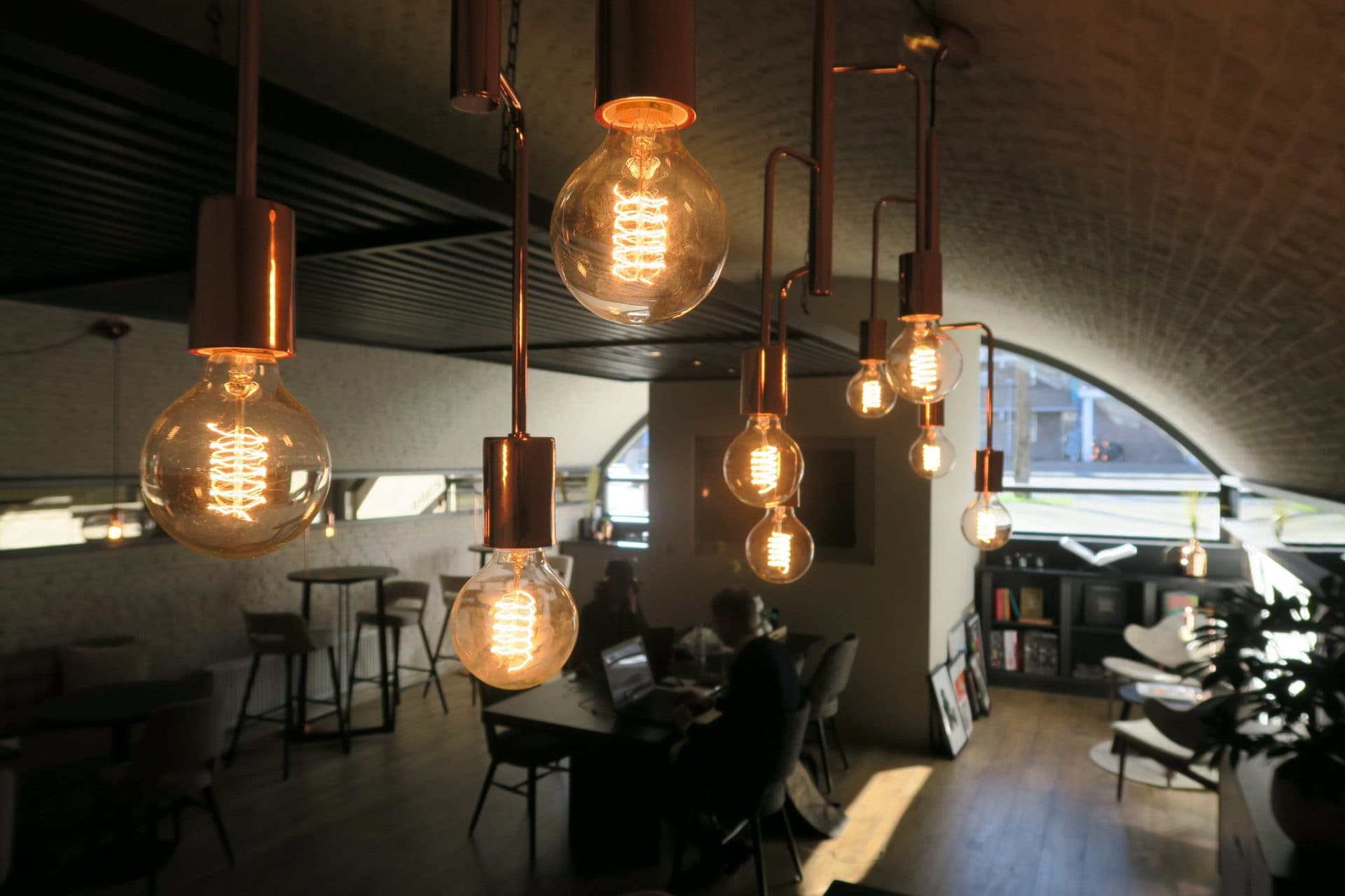 Koperen hanglampen met gloeidraadlampen onder de boog, terwijl mensen aan de grote tafel op hun laptop werken, met een boekenkast en loungestoelen bij het boograam