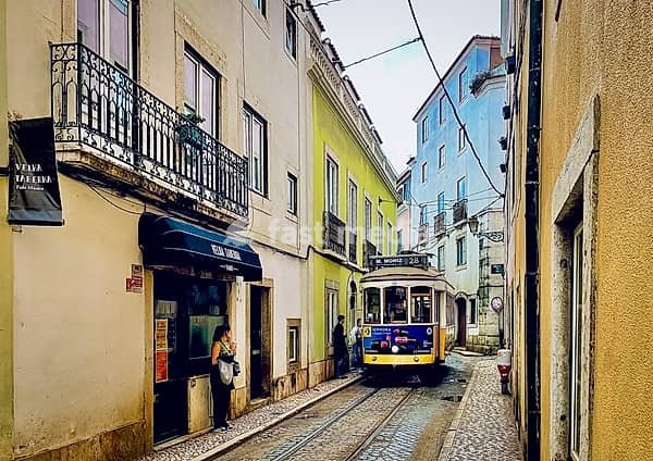 Charming scene from Lisbon's old town with the famous tram line 28, which snakes through a narrow, colorful alley. Colourful house facades in yellow, blue and ochre with traditional balconies