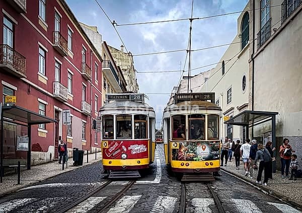 Lively scene from Lisbon with two traditional yellow trams on line 28 that travel through a narrow street between colorful buildings. People wait at the bus stop while the old trams shape the typical cityscape