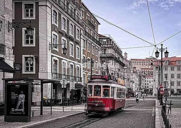 City view of a classic red tram in Lisbon, Portugal. The scene features historic buildings with typical Portuguese architecture, cobblestone streets and European-style lanterns. The sky is slightly cloudy and passers-by enliven the scene