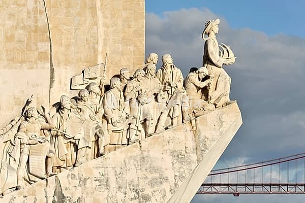 Monument of Discoveries, Padrao dos Descobrimentos, monument with important figures of Portuguese seafaring on the banks of the Tagus River, Belem, Lisbon, Portugal, stone reliefs and figures on a monument in front of a red bridge and a cloudy sky, Lisbon, Portugal