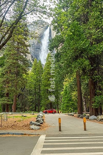 Lower yosemite fall cascades down a granite cliff into a lush green valley, framed by tall trees and a walkway with a parked red car near the scenic natural attraction