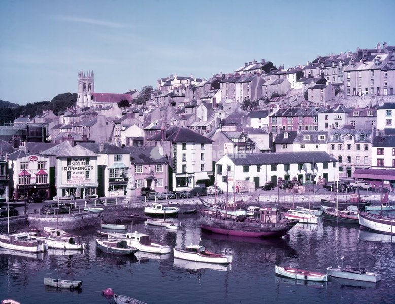 Brixham, Devon, c1955. Town built around a natural harbour, which in addition to leisure craft, provides anchorage for what is now one of England's largest remaining commercial fishing fleets. View showing the Strand Baker cafe; Kaye's yacht supplies, ironmonger and plumber; the Ship cafe; and the…