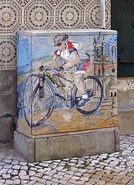 Street art painting of cyclist on an electrical box with traditional Portuguese tiles and cobblestones