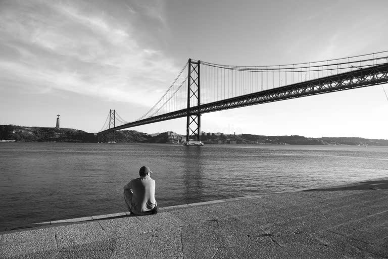 Ein Mann sitzt am Tejo, in der Nähe der Brücke, die sich im Abendlicht über das Wasser spannt