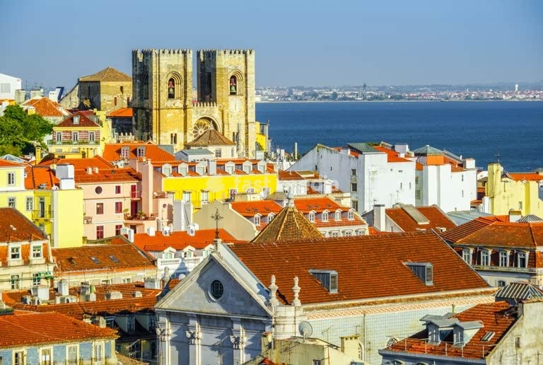 Colorful rooftops of Lisbon's Alfama district with the historic Sé Cathedral and a cargo ship on the Tagus River under a bright blue sky.