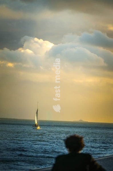 Solitary figure watches sailboat departing into golden Tagus estuary at dusk, Lisbon, Portugal