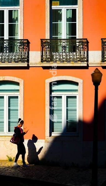 Woman on smartphone cast in dramatic shadow against vivid orange facade, Alcântara, Lisbon, Portugal
