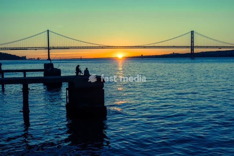 Fishermen silhouetted at sunset before the 25 de Abril Bridge, Tagus River, Lisbon, Portugal