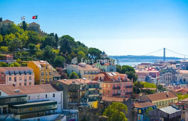 Morning panorama over Lisbon's colorful hillside houses with flags on the castle walls and Tagus River bridge under clear blue sky in Portugal