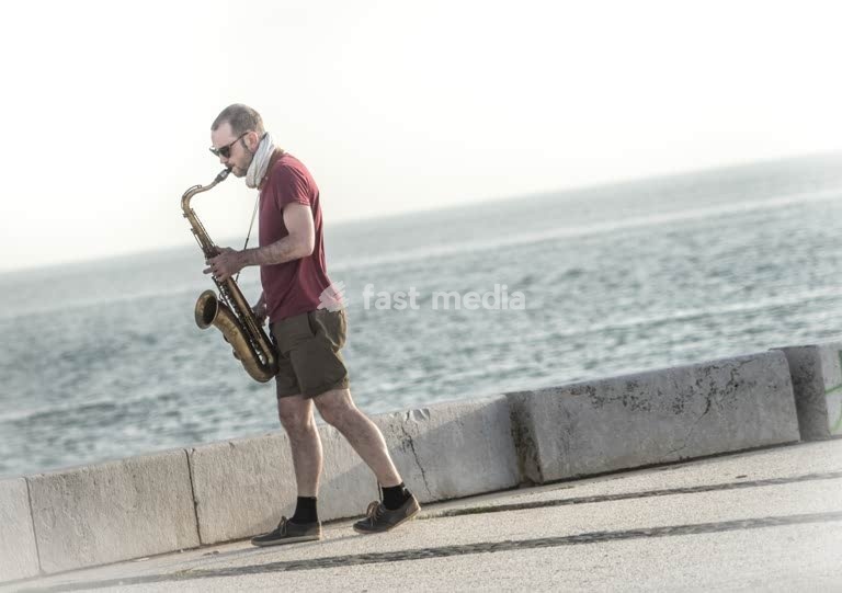 Street musician playing saxophone on Lisbon waterfront promenade with view on River Tagus, Portugal - casual performance by the river