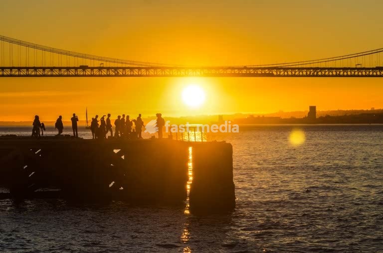 Crowd watching sunset silhouetted on pier, 25 de Abril Bridge, Lisbon, golden evening scene