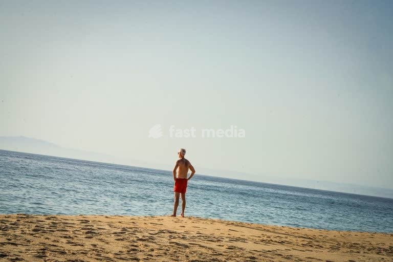 Senior man standing alone on a quiet beach, gazing at the sea—capturing a moment of solitude, reflection, and peaceful coastal ambience