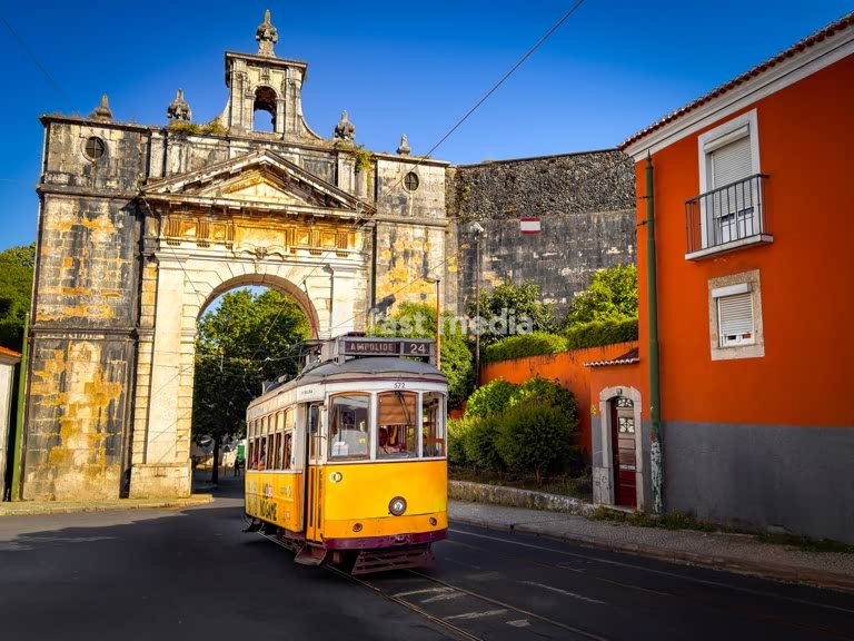 Historic yellow Lisbon tram 24E passes through an archway of the ancient aqueduct with colorful Portuguese buildings under bright blue sky in Portugal