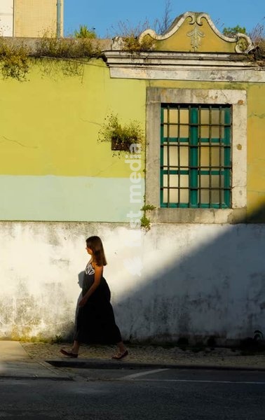 Woman walks past weathered yellow and white wall with green shuttered window casting dramatic shadows on cobblestone street in old Lisbon