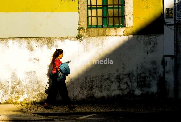 Woman walks past weathered yellow and white wall with green shuttered window casting dramatic shadows on cobblestone street in old Lisbon
