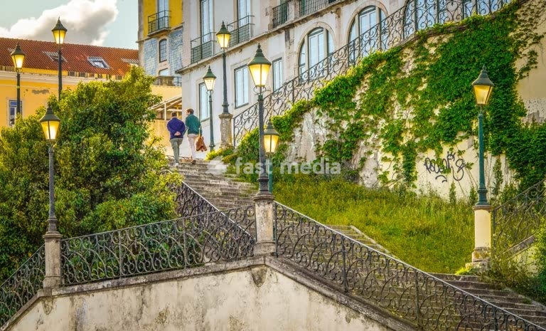 A couple climbing up sunlit stairs in Lapa, Lisbon, surrounded by vintage lamps, lush greenery and glowing rays of golden hour light