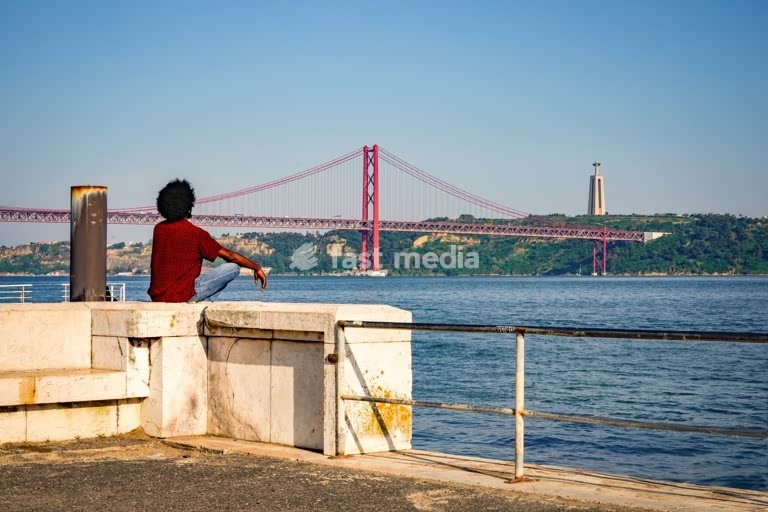 Young man relaxing by the Tagus River in Lisbon, facing the 25th April Bridge and Cristo Rei monument on a clear, sunny day