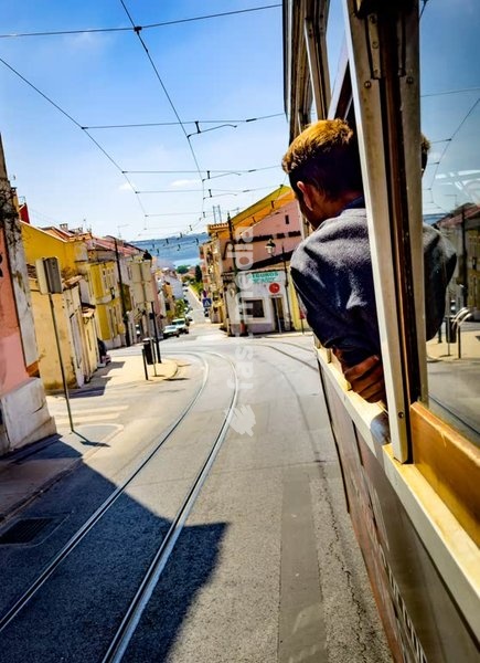 Young man looking out of vintage tram in Lisbon's Boa-Hora district on a sunny day, with view of colorful houses and the Tagus River ahead