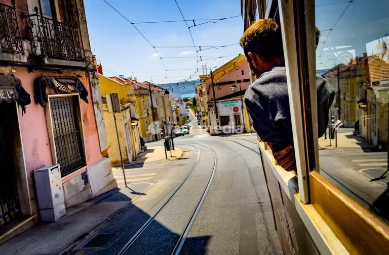 Secret Lisbon: Young man looking out of vintage tram in Lisbon's Boa-Hora district on a sunny day, with view of colorful houses and the Tagus River