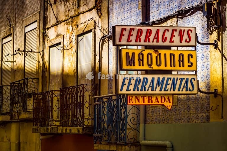 Vintage neon shop sign for hardware and machines on a weathered building facade in Lisbon, Portugal, with azulejos tiles and iron balconies