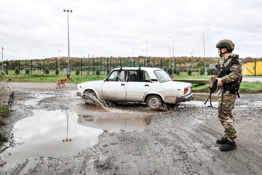 An armed Zaporizhzhian police officer stands in the street as a car drives through a puddle during a humanitarian evacuation mission for a family, including an elderly woman with limited mobility, from the settlement of Komyshuvakha, Zaporizhzhia region, Ukraine, October 19, 2026