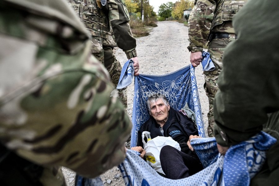 Zaporizhzhia police officers and a family member carry an elderly bedridden woman in a blanket towards a car during an evacuation from the front-line settlement of Komyshuvakha, Zaporizhzhia region, Ukraine, October 19, 2026