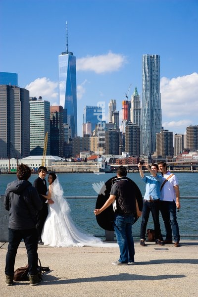 USA, New York, Brooklyn Bridge Park, two young men taking a selfie with the Lower Manhattan skyscraper skyline as a backdrop beside a couple having their wedding photograph taken on the Fulton Ferry Landing