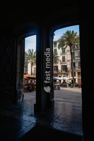 Spain, Balearic Islands, Majorca, Palma de Mallorca, Old Town. View into Placa de La Llotja from inside the medieval guild hall. People at restaurant table under umbrellas in the old market square