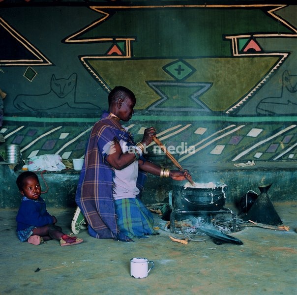 Anna Kabin with her granddaughter Sbongile of Ramokgeletsane, KwaNdebele, preparing the Ndebele staple diet of maize meal and sour milk. The cooking hut is roofed with corrugated iron sheeting; the only outlet for smoke is a tiny window. Above a low mud bench running the length of the room is a…