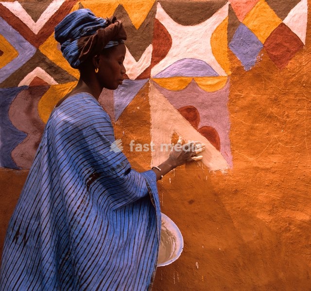 Silla Camara applies fo khoule - a paste of ground white limestone and water - to the mud wall, tagaye, of her house. Traditionally, only red earth, fo doumbe, black, fo bine, and white were used. In more recent years, the younger generation of women have added washing blue, boula; yellow, makka;…