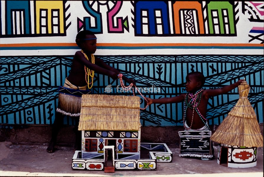 Ndebele children, dressed in traditional beaded attire, play with models of Ndebele thatched homes. Behind them, a painted mural can be seen