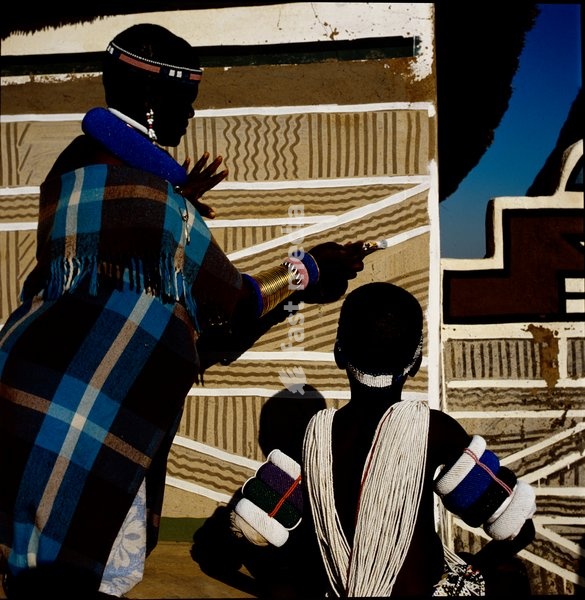 An Ndebele woman and child, wearing traditional attire, painting a wall in graphic geometric patterns