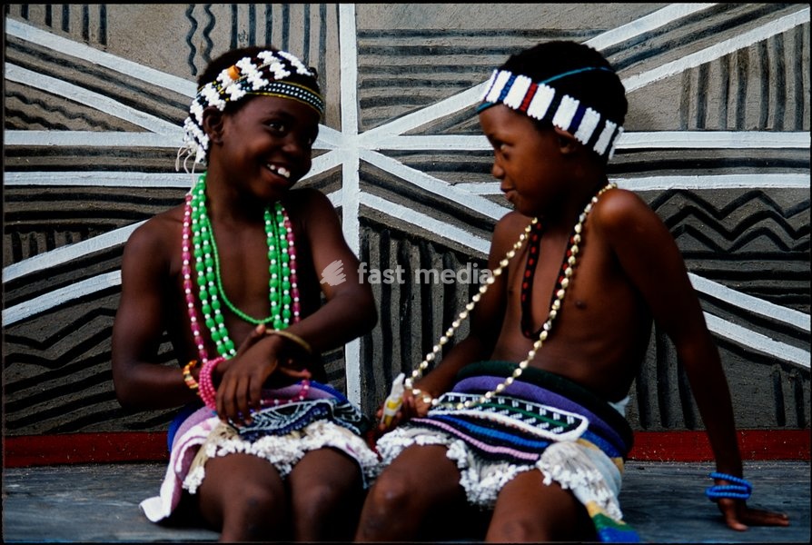 Ndebele children, wearing beaded jewellery, aprons and headbands, pose in front of a painted wall