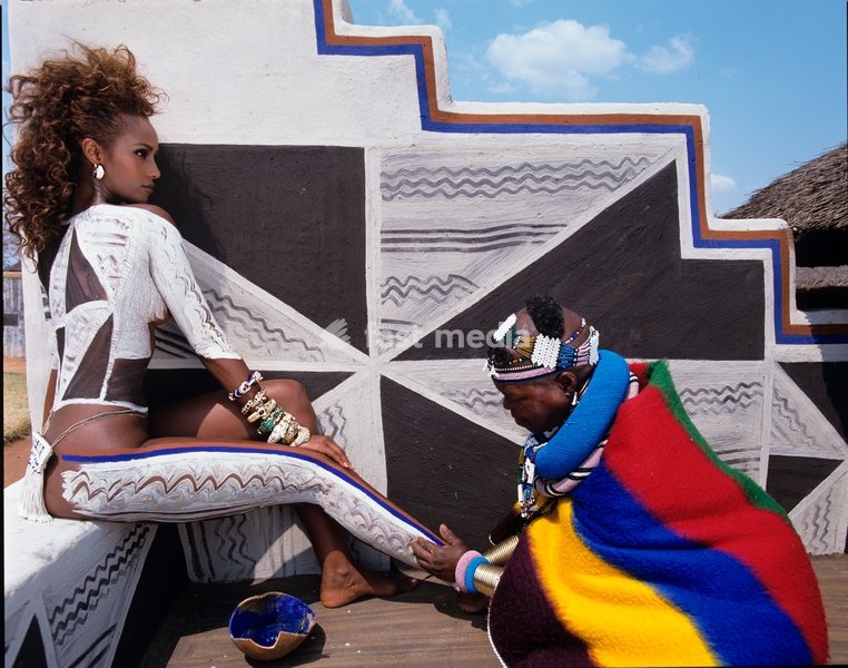 An Ndebele woman applies body paint to a model in front of a wall painted in geometric patterns