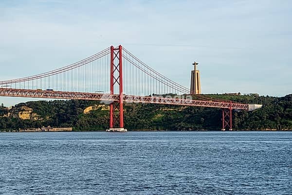 Red bridge over a river, next to it a large statue on a wooded hill, Ponte 25 de Abril, Lisbon, Portugal, Europe