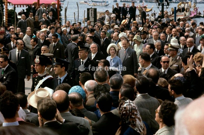 Queen Elizabeth II of the United Kingdom is visiting Venice for the first time, walking along Piazza San Marco, densely crowded by the venetians; the young Queen, visiting Italy for the centenary of the national unification, is guided by Mayor Giovanni Favaretto Fisca, at her right side. Venice…