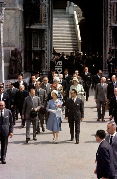 Queen Elizabeth II of the United Kingdom, with a flower in hand, walks along Piazza San Marco, in front of the Porta della Carta, beside her guide and closely followed by the Prince Philip; the royal couple is in Italy for the centenary of the national Unification, and their visit attracted a large…