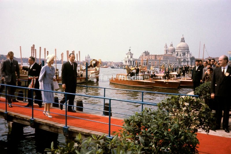 Queen Elizabeth II of the United Kingdom, visiting Venice for the first time, in the occasion of the celebrations for the national unification's centenary, crosses the red footbridge, followed by the Mayor Giovanni Favaretto Fisca, with the tricolor band and by her husband, Prince Philip, just got…