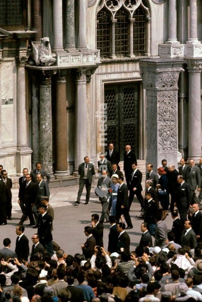 Queen Elizabeth II of the United Kingdom, with a flower in hand, walks along Piazza San Marco beside her guide and closely followed by the Prince Philip; the royal couple is in Italy for the centenary of the national Unification, and their visit attracted a large crowd in every city they touched.…