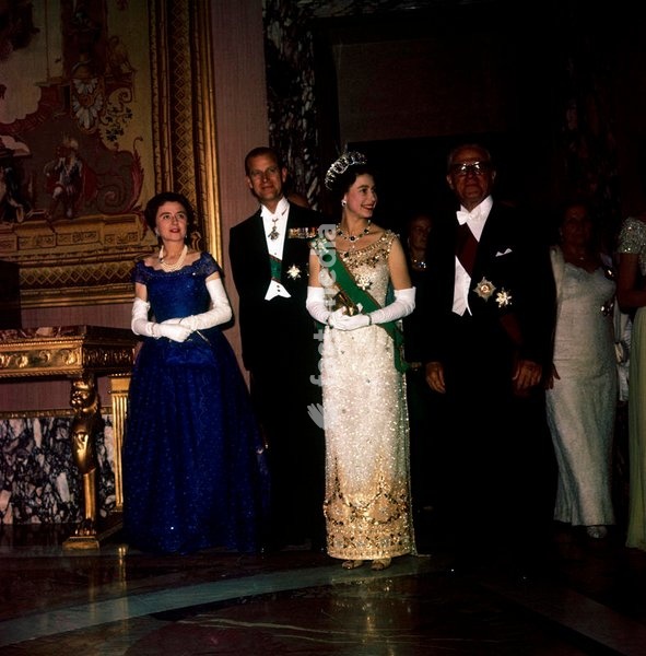 The Queen of the United Kngdom, in a sequins tulle dress, stands in a hall of the Quirinal Palace, between the italian President of the Republic Giovanni Gronchi and her husband, the Prince Philip of Edimburgh; on the left, in a blue dress, is the italian first lady Carla Bissatini Gronchi;…