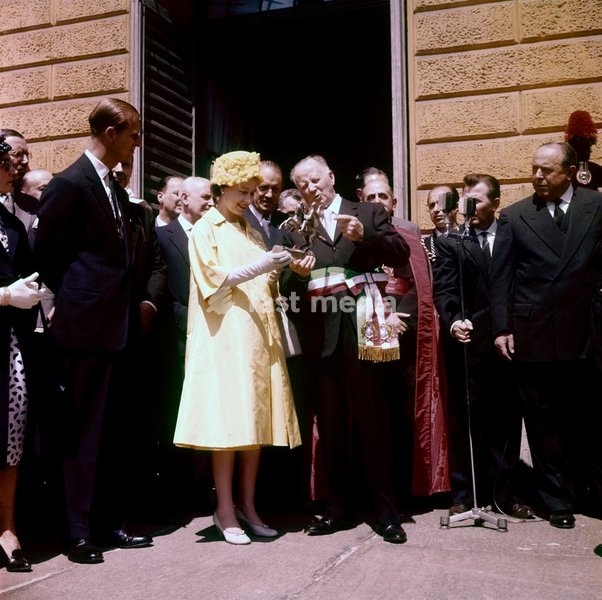 Queen Elizabeth, dressed in yellow and accompanied by her husband, the Prince Philip, Duke of Edimburgh, holds a bronze statuette received by the Mayor of Castiglione Torinese, welcoming the royal couple in their visit in Italy for the centenary of the Unification; the gift commemorates the 126…