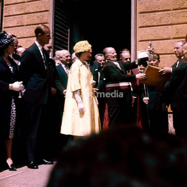 Queen Elizabeth, dressed in yellow and accompanied by her husband, the Prince Philip, Duke of Edimburgh, receives the welcome of the Mayor of Castiglione Torinese; the mayor present them some gifts, a book and a bronze statuette, in the memory of 126 british prisoners preserved by his fellow…