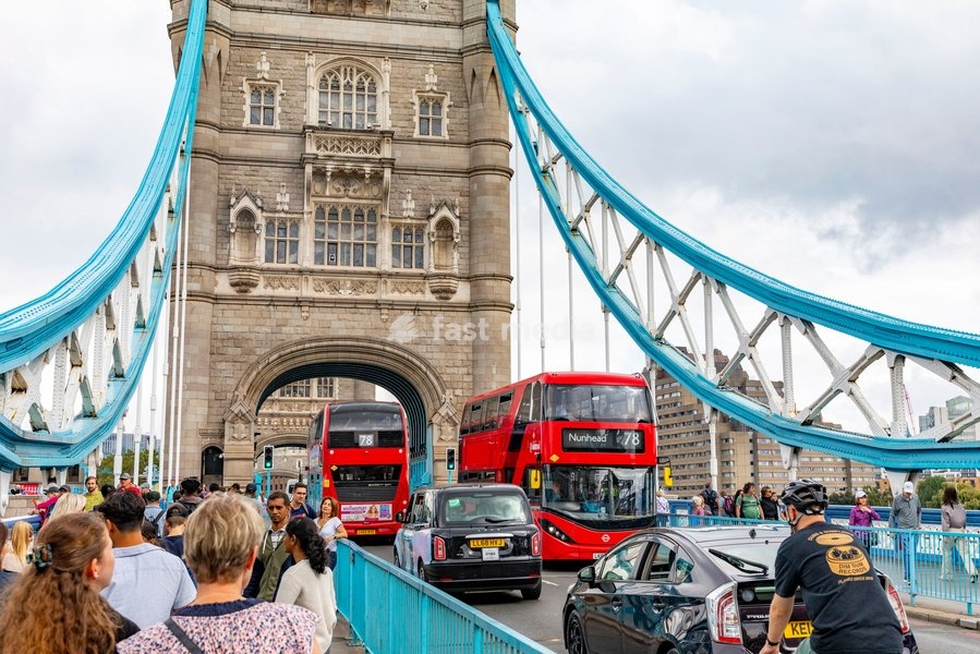 Red double decker London buses travel across Tower Bridge whilst visitors walk alongside the traffic